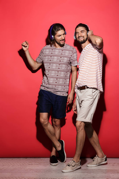 Full length portrait of a two young cheerful twin brothers isolated over red background, listening to music with headphones - Foto, immagini