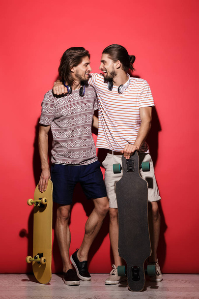 Full length portrait of a two young smiling twin brothers isolated over red background, listening to music with headphones, holding skateboards - Foto, Imagem