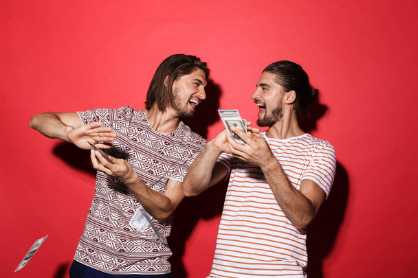 Portrait of a two young cheerful twin brothers isolated over red background, throwing out money banknotes - Photo, Image
