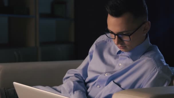 Ένας άνδρας κάθεται σε ένα φορητό υπολογιστή σε ένα γραφείο σκούρο. Ο άνθρωπος που λειτουργεί μέχρι αργά το βράδυ - Πλάνα, βίντεο