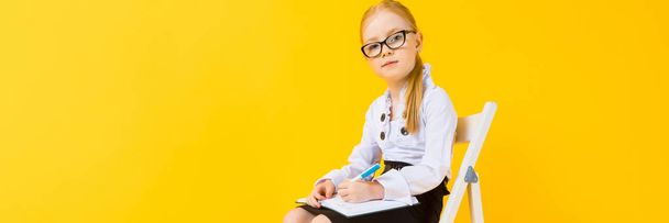 Mädchen mit roten Haaren auf gelbem Hintergrund. ein charmantes Mädchen mit durchsichtiger Brille sitzt auf einem weißen Stuhl und macht Notizen in einem Notizbuch. - Foto, Bild