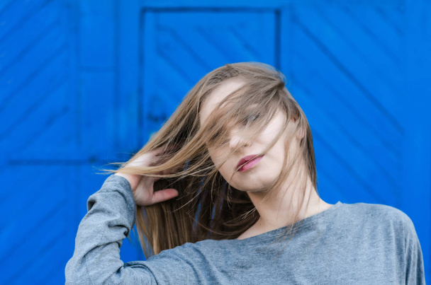 Attractive girl with long blond hair fluttering in the wind on a blue background. Youth sexuality. Independent girl. Dynamic portrait. - Fotografie, Obrázek