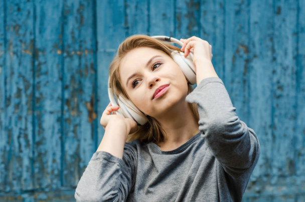 Cute girl listening to music in white wireless headphones on the background of the old board. Electronic music. Musical style. Pleasant sounds. The pleasure of sound. - Foto, afbeelding