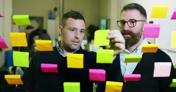 Two creative and thoughtful in a creative agency with colored and planning notes hanging from the glass and behind the working people. Concept: team work, agency, creative, business - Materiał filmowy, wideo