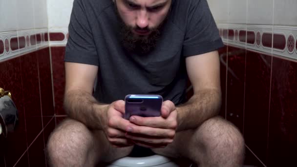 lähikuva nuoresta valkoihoisesta miehestä istumassa vessassa lähettämässä viestiä älypuhelimellaan. Lähikuva nuori valkoihoinen mies käyttää älypuhelinta kylpyhuoneessa
. - Materiaali, video