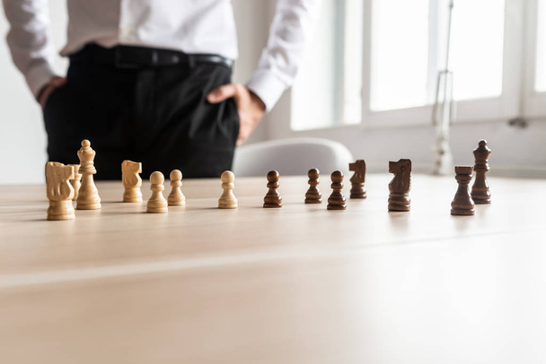 Businessman standing at his office desk looking at black and white chess pieces arranged on the wooden desk. With copy space on the lower part of the image. - Fotó, kép