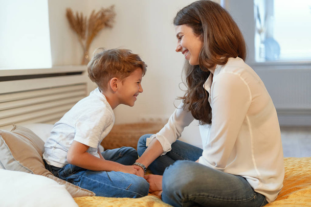 Mom and son smiling, looking at each other, having fun, spend time together in a relaxing atmosphere at home while sitting - Foto, imagen