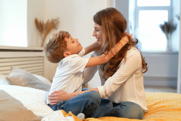 Mãe e filho sorrindo, abraçando, olhando um para o outro, se divertindo em uma atmosfera relaxante em casa enquanto sentados
 - Foto, Imagem