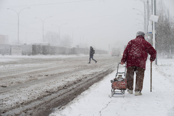 Una tormenta de nieve en la ciudad. Una anciana con un bastón y una bolsa sobre ruedas recorre el camino durante una tormenta de nieve. Enfoque selectivo
. - Foto, imagen