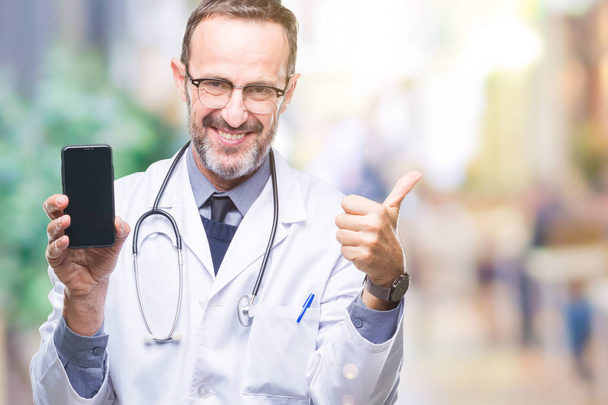 Middle age senior hoary doctor man showing smartphone screen over isolated background happy with big smile doing ok sign, thumb up with fingers, excellent sign - Photo, Image