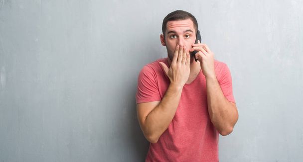 Молодий кавказький чоловік над сірою гранжевою стіною, що говорить по телефону, прикриває рот рукою, шокованою соромом за помилку, вираження страху, наляканий тишею, таємна концепція
 - Фото, зображення