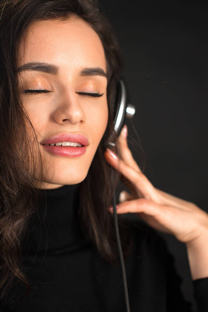 Hermosa mujer joven disfrutando escuchando la música en auriculares inalámbricos con los ojos cerrados sobre fondo negro oscuro. Primer plano.
. - Foto, Imagen