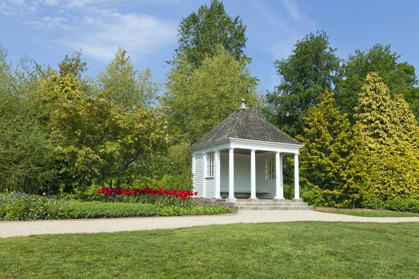 Casa de verano blanca junto a un sendero en un jardín de primavera con tulipanes rojos, arbustos y árboles
 . - Foto, Imagen