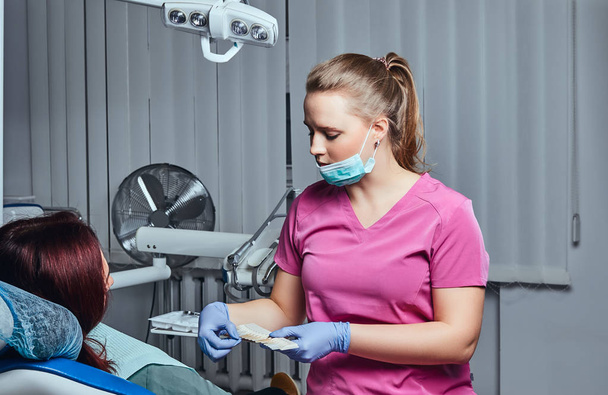 Una mujer dentista comprueba el nivel de blanqueamiento dental con un color de dentista en el consultorio del dentista
. - Foto, Imagen