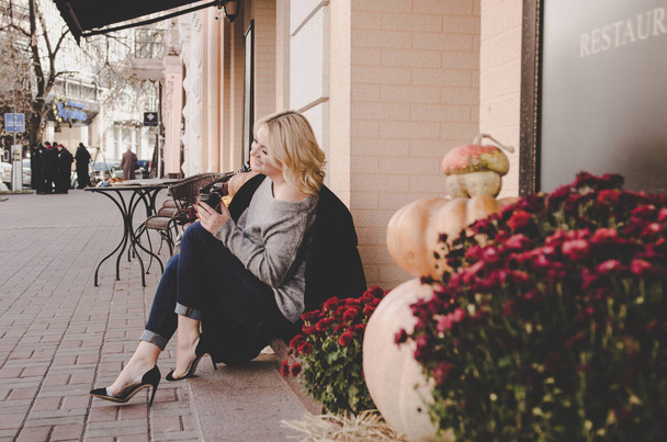 young blonde woman in black jacket sitting near pumpkins and drinking coffee  - Foto, Imagen