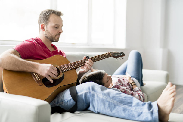 Jovem casal sentado no sofá na sala de estar e tocando guitarra
 - Foto, Imagem