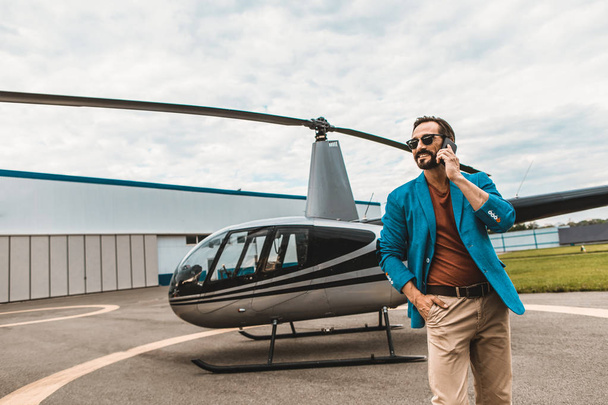 Attractive man in colorful clothes standing near the helicopter with smartphone - Zdjęcie, obraz