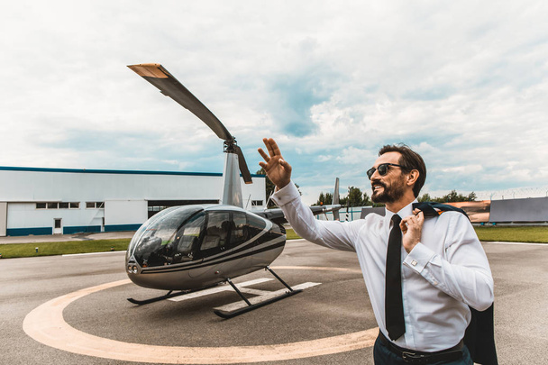 Cheerful man putting one hand up while standing near the helicopter - 写真・画像