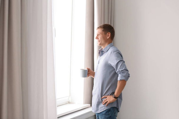 Mature man with cup of drink near window with open curtains at home. Space for text - Foto, Imagen