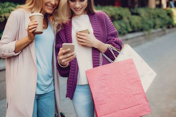 Cropped portrait of beautiful middle-aged woman and her daughter looking at cellphone and smiling. They holding cups of coffee and shopping bags - Foto, imagen