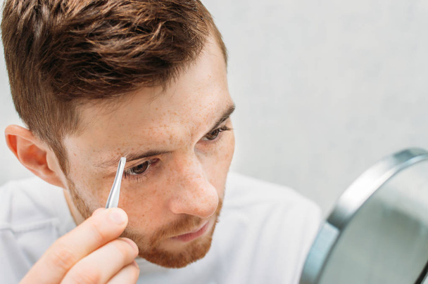 young man plucks his eyebrows in front of a mirror. Closeup portrait - Fotoğraf, Görsel