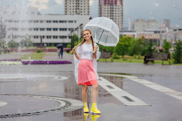 Genç güzel kız sarı çizmeli iki örgüler ve şeffaf şemsiye ile Çeşme duruyor. Şehirde yağmurlu gün. - Fotoğraf, Görsel