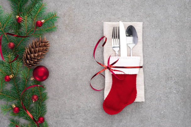flat lay with fork, spoon with knife in santa sock wrapped by red ribbon on table with pine cone and branch decorated by christmas balls  - Photo, Image
