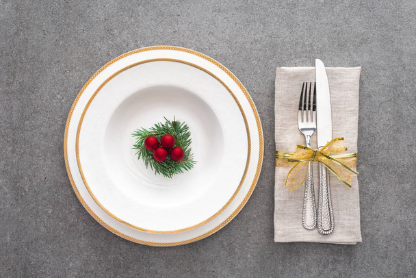 view from above of served plate with decorated evergreen branch and christmas balls near fork with knife wrapped by ribbon on grey surface - Фото, изображение
