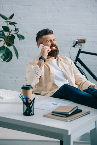 Smiling bearded businessman sitting in modern office and talking on smartphone - Foto, imagen