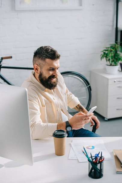 Usmíval se vousatý podnikatel sedí u stolu a při pohledu na smartphone v moderní kanceláři  - Fotografie, Obrázek