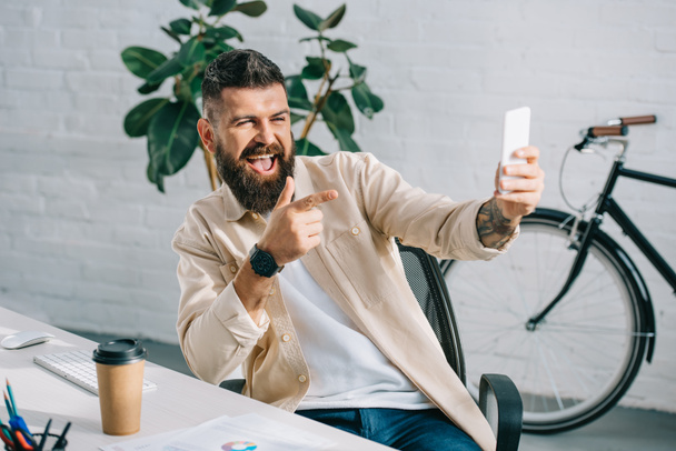 bärtiger Geschäftsmann zeigt beim Selfie im Bürostuhl in die Kamera - Foto, Bild