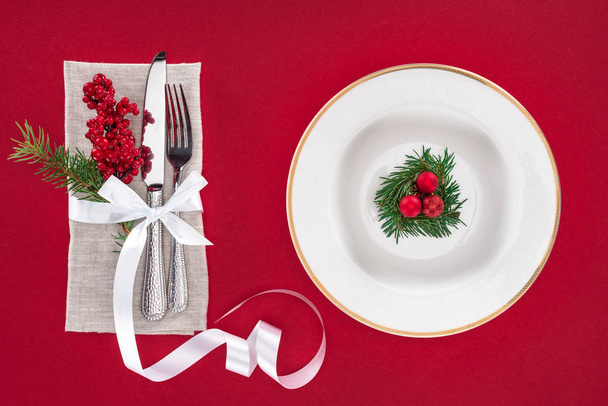 view from above of fork, knife, red berries wrapped by ribbon and plate with evergreen tree branch with christmas balls isolated on red - Fotografie, Obrázek