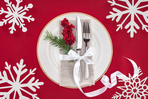 view from above of fork, knife, red berries wrapped by ribbon surrounded by decorated snowflakes isolated on red - Фото, изображение