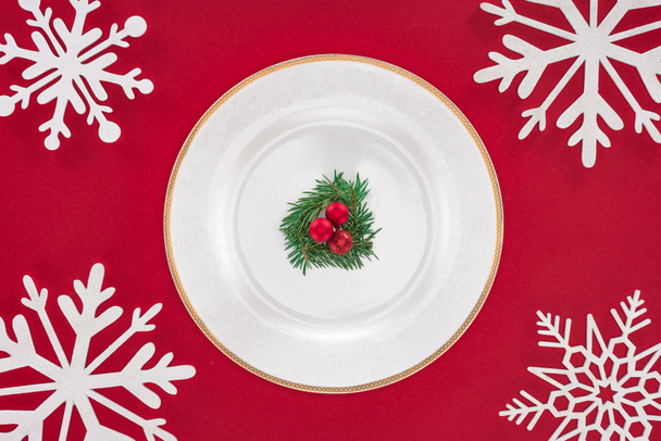 vista superior de la placa con rama de árbol siempreverde y bolas de Navidad en el plato rodeado de copos de nieve aislados en rojo
 - Foto, Imagen