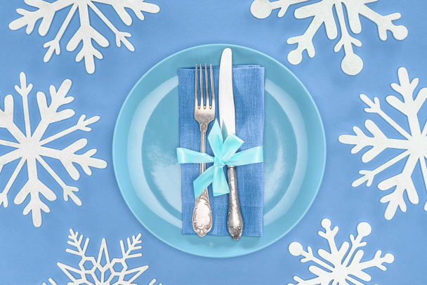 verhoogde uitzicht op vork en mes verpakt door feestelijk lint op plaat omgeven door sneeuwvlokken geïsoleerd op blauw - Foto, afbeelding