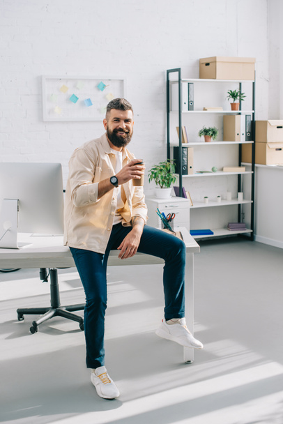 Smiling businessman sitting on office desk and holding disposable cup - Foto, Bild
