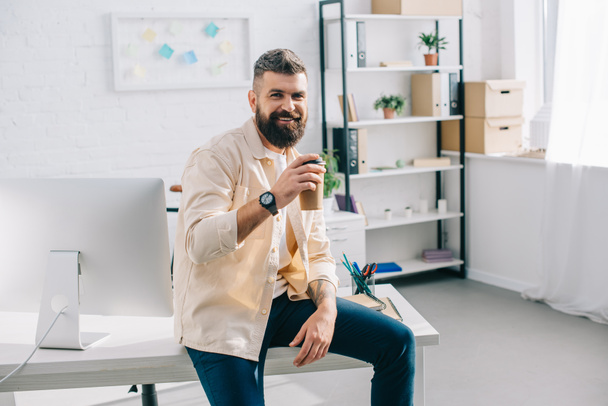 Улыбающийся бородатый бизнесмен сидит на рабочем столе с бумажной чашкой
 - Фото, изображение