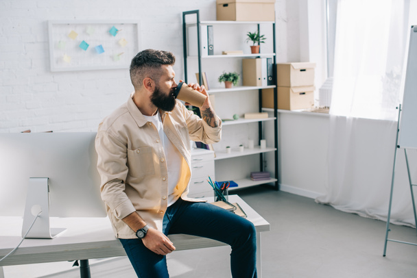 parrakas liikemies istuu toimistopöydällä ja juo kahvia kertakäyttökupista
  - Valokuva, kuva