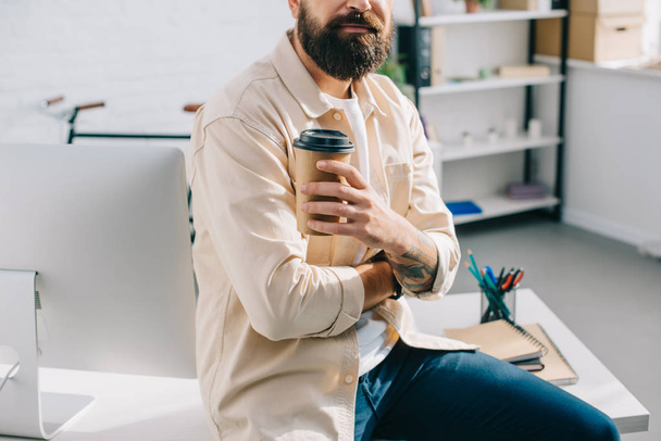 abgeschnittene Ansicht eines bärtigen Mannes, der auf einem Schreibtisch sitzt und eine Kaffeetasse aus Papier hält - Foto, Bild