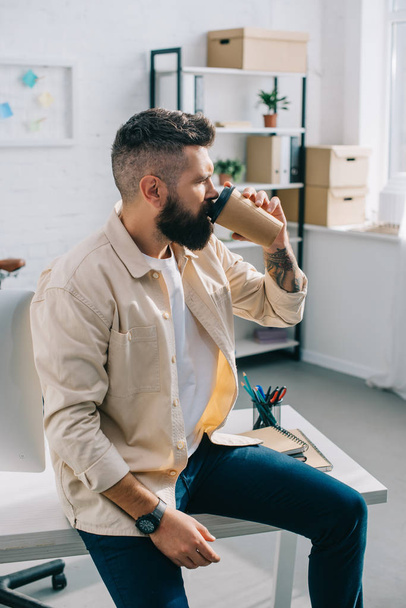 bearded businessman sitting on desk and drinking coffee from paper cup in office - Foto, imagen