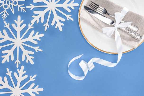top view of fork and knife wrapped by festive ribbon on plate surrounded by snowflakes isolated on blue - Photo, Image