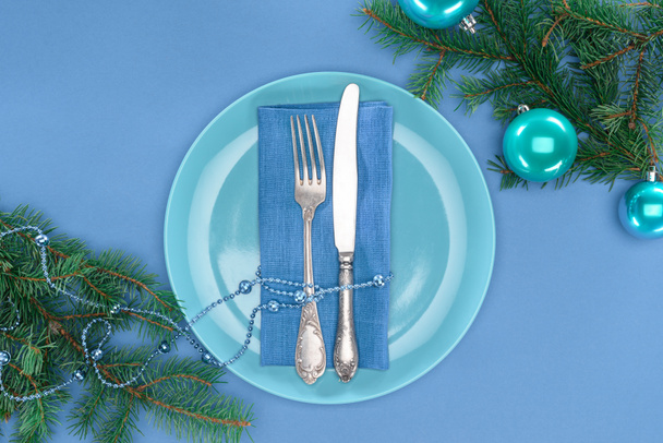 verhoogde uitzicht op vork en mes door parels op plaat in de buurt van groenblijvende takken omwikkeld met kerstballen geïsoleerd op blauw - Foto, afbeelding