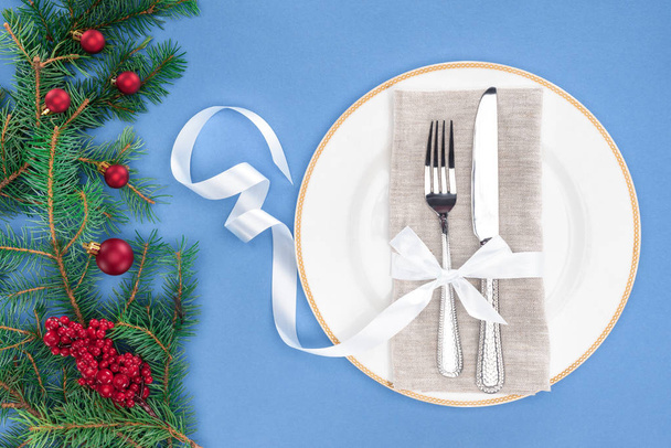top view of fork and knife wrapped by ribbon on plate near evergreen branches with christmas balls and red berries isolated on blue - Foto, Imagen