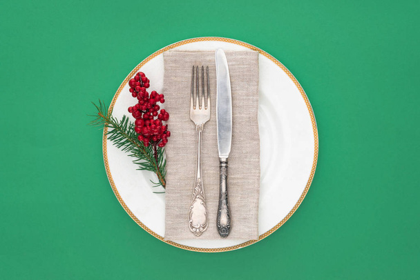 верхний вид плиты с вилкой, ножом, вечнозеленой веткой и красными ягодами на табличке изолированы на зеленый
  - Фото, изображение