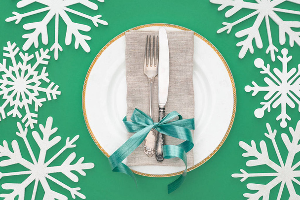 top view of plate with fork and knife wrapped by festive ribbon surrounded by snowflakes isolated on green - Fotografie, Obrázek