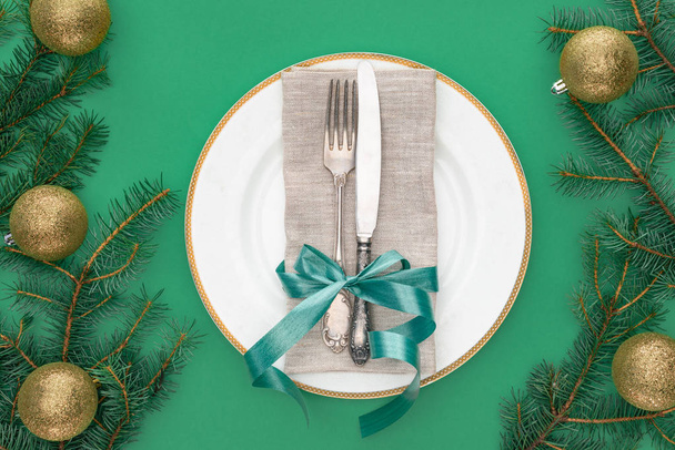 вид сверху вилки и ножа, завернутый праздничной лентой на тарелке в окружении вечнозеленых ветвей деревьев с рождественскими шарами, изолированными на зеленом
 - Фото, изображение