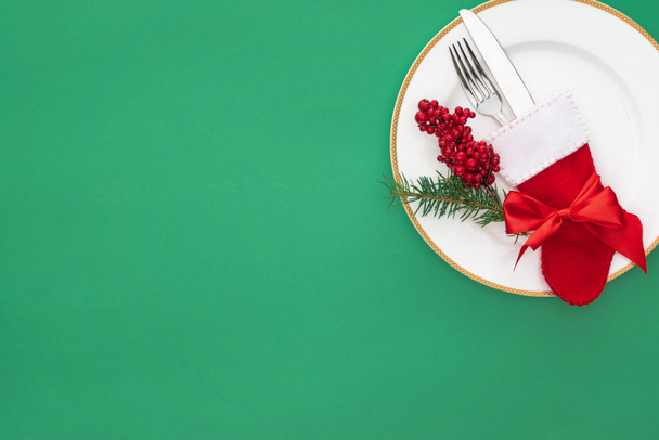 elevated view of red berries, evergreen tree branch and fork with knife in christmas sock on plate isolated on green - Photo, Image