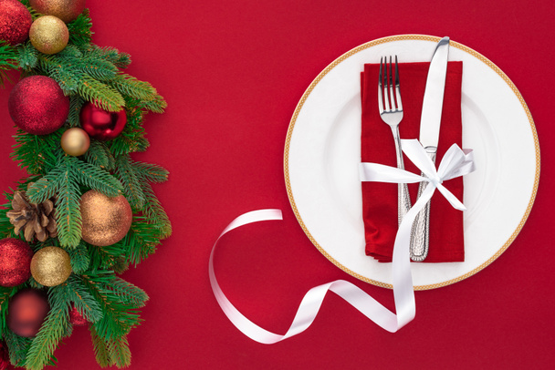 top view of fork and knife wrapped by festive ribbon on plate near evergreen tree branches with christmas balls isolated on red - Valokuva, kuva