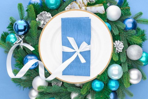 elevated view of napkin wrapped by festive ribbon on plate surrounded by evergreen tree branches with christmas balls isolated on blue - Photo, Image