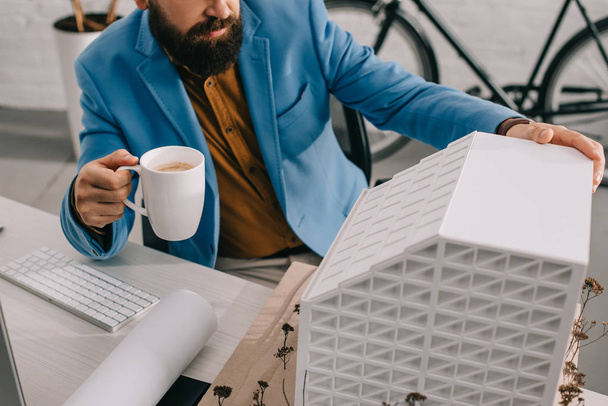 abgeschnittene Ansicht eines erwachsenen männlichen Architekten in formaler Kleidung, der eine Tasse Kaffee hält und im Büro an einem Projekt arbeitet - Foto, Bild